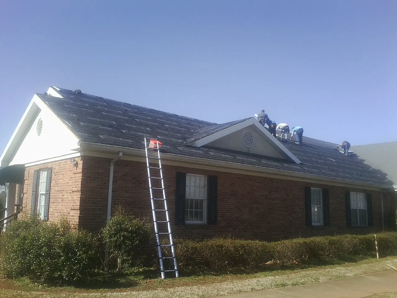 Crew tearing shingles off a brick ranch roof with a ladder set — Emergency Roof Repair work by our crew in Lapeer