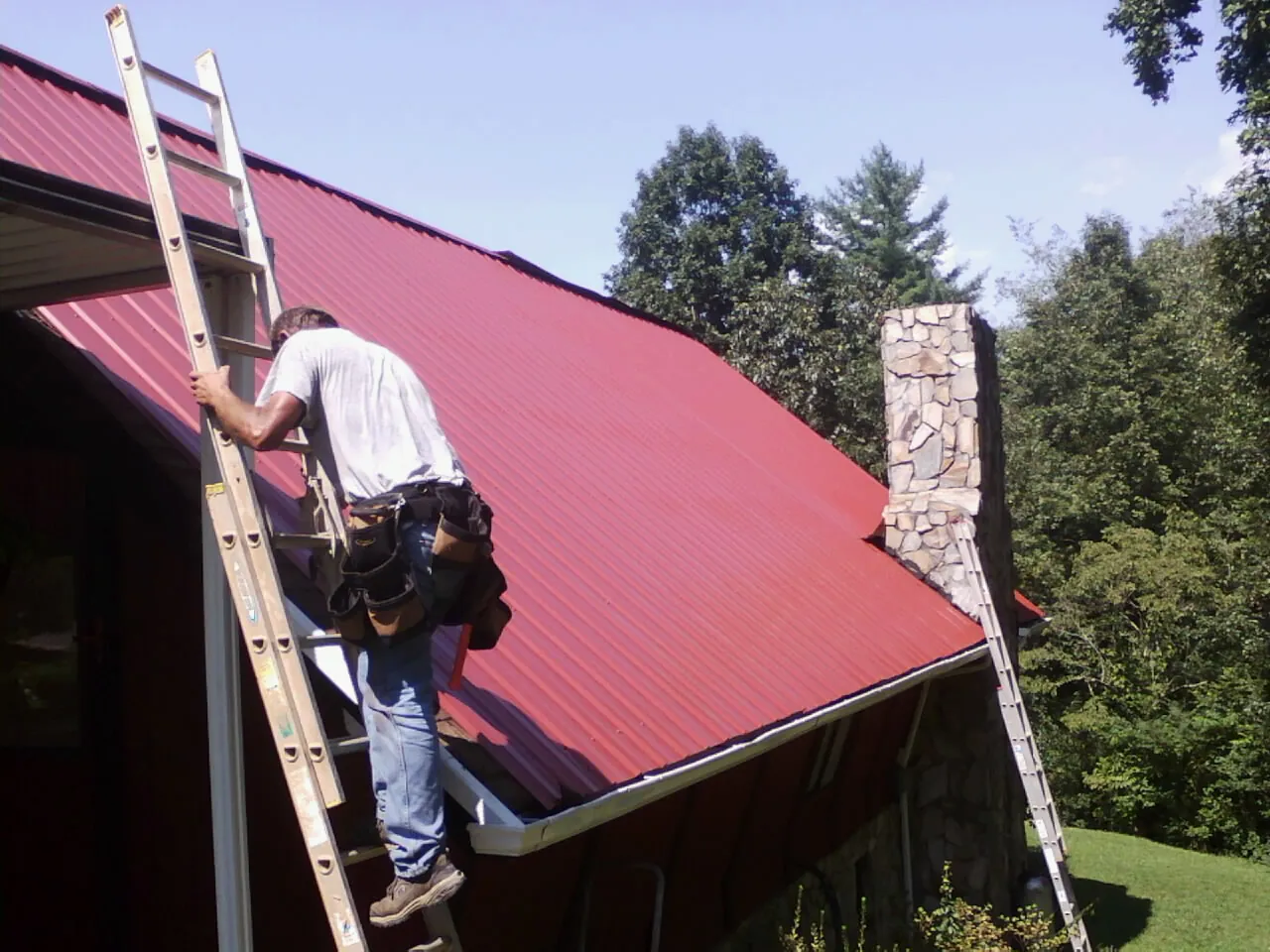 Roofer on a ladder beside a red metal roof and stone chimney — Soffit & Fascia Repair by Roofing Brothers in Lapeer